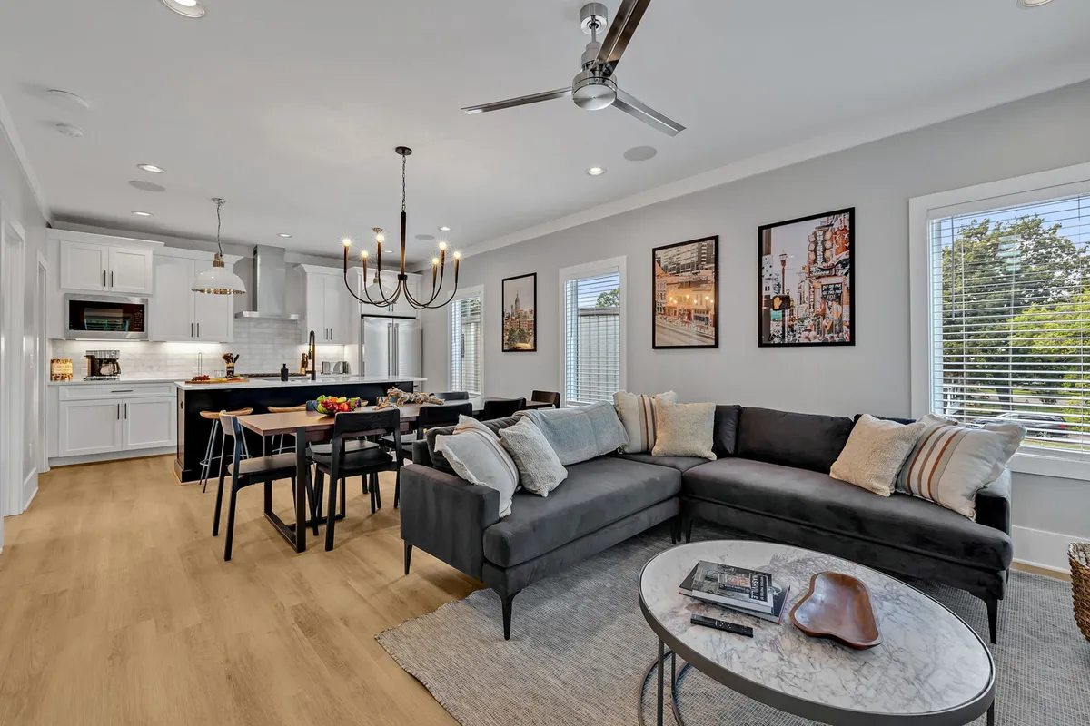 The image showcases a stylish, open-concept living space featuring a plush sofa, chic dining table beneath a statement chandelier, and a sleek kitchen island—perfect for gatherings and entertaining. The setting is modern yet inviting, with warm lighting and thoughtful details that create a sense of comfort and connection.