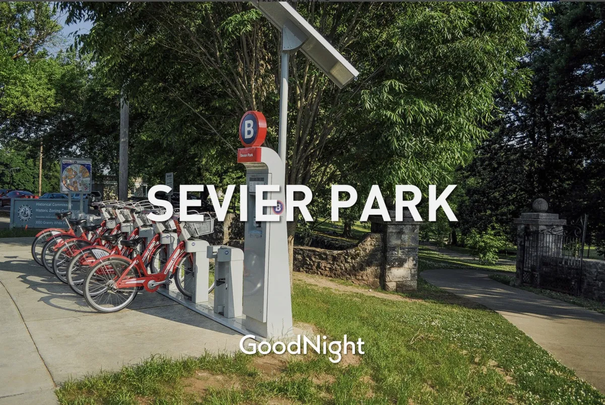 A welcoming park entrance bordered by trees with a nearby bike rental station. The scene suggests opportunities for outdoor activities, including biking, walking, picnicking, and community events, all in a vibrant, green setting.