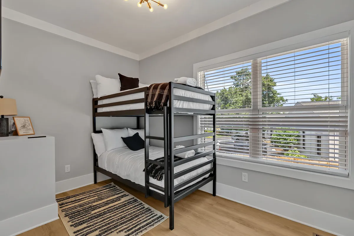 A cheerful bunk room features a sturdy bunk bed, a soft rug for play or reading, and a window allowing daylight to filter in. The space feels playful and welcoming, catering to younger guests or groups seeking a cozy sleepover vibe.