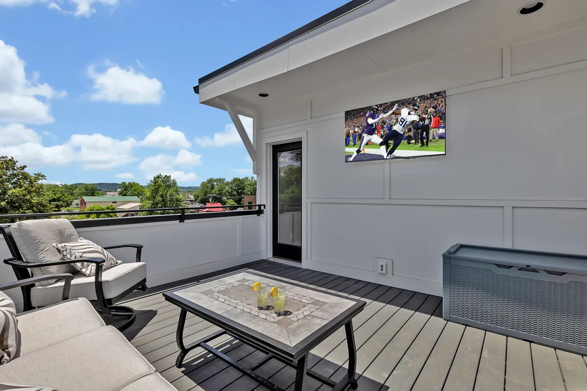 A modern outdoor lounge area with comfortable seating and a television mounted on the wall, overlooking an open landscape. The setup is designed for socializing, with the TV making it ideal for group events or game nights. The space feels open and exclusive, emphasizing luxury and entertainment.