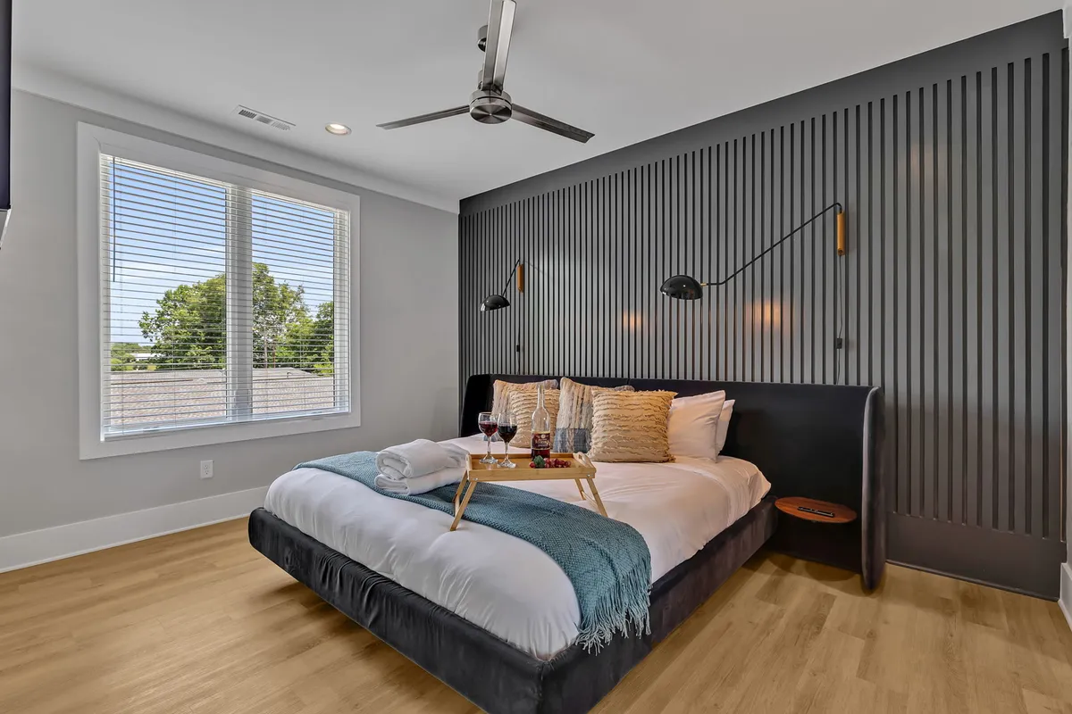 A king bed with decorative pillows sits beneath a textured accent wall, flanked by a bedside table with fruits and glasses. Large windows let in abundant natural light, creating a serene, inviting atmosphere ideal for relaxation. The overall decor feels luxurious and tranquil, reinforcing the sense of a private sanctuary.