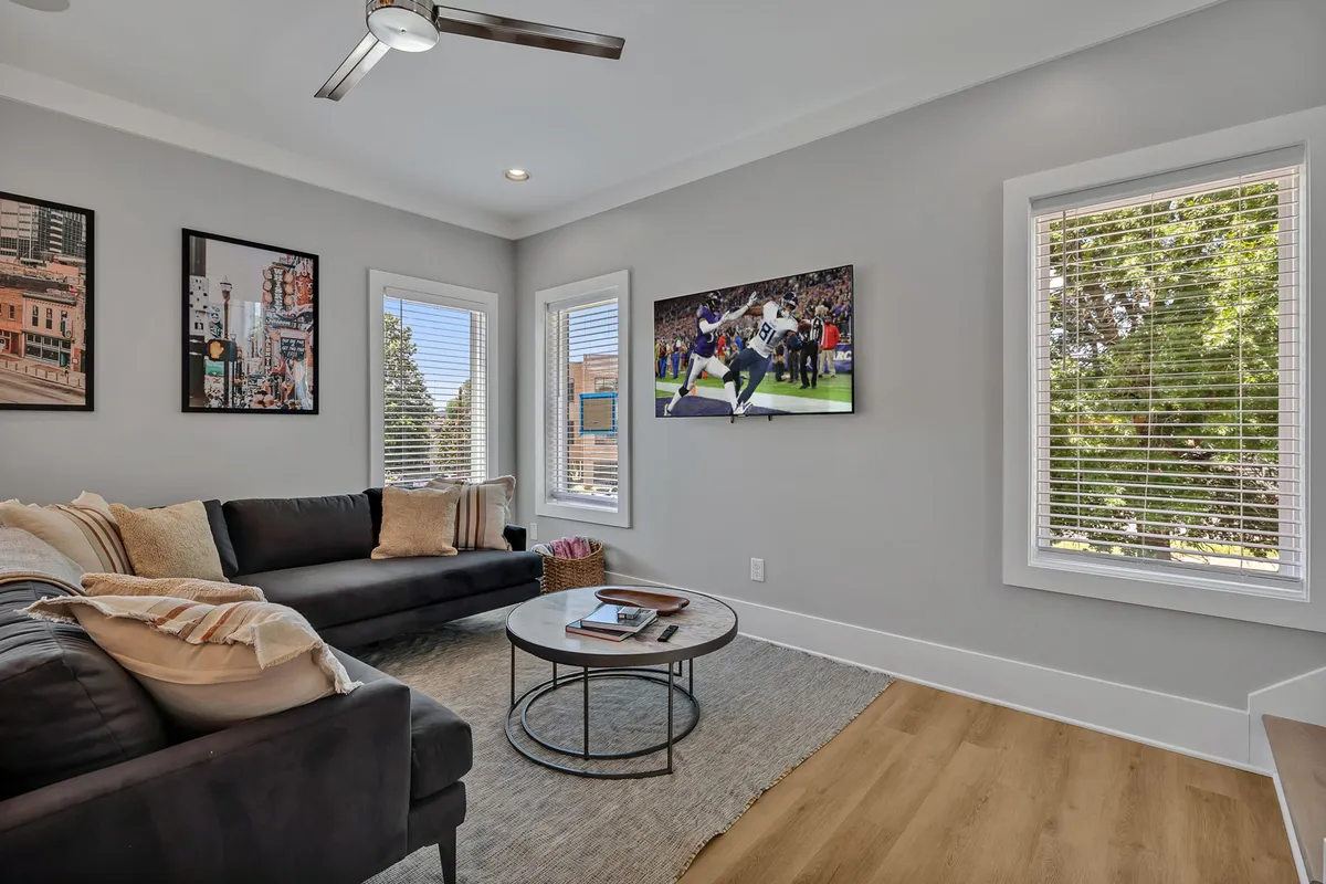 A bright, open living room with a plush sectional sofa, a modern coffee table, and a large Smart TV. Natural light streams through expansive windows, and stylish framed artwork adorns the walls. The space looks inviting for gatherings, movie nights, or relaxing downtime.